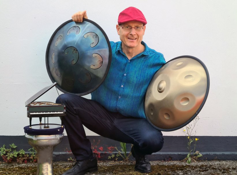 Ein Mann mit roter M&uuml;tze h&auml;lt zwei Handpans und sitzt neben einem kleinen Klaviermodell auf einer Trommel. Er tr&auml;gt ein blaues Hemd und l&auml;chelt in die Kamera., &copy; Holger Mantey