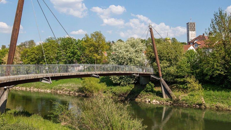 Eine moderne Fußgängerbrücke überspannt einen Fluss, umgeben von grüner Vegetation. Im Hintergrund ist ein Kirchturm zu sehen., © Stuttgart-Marketing GmbH Eine moderne Fußgängerbrücke überspannt einen Fluss, umgeben von grüner Vegetation. Im Hintergrund ist ein Kirchturm zu sehen., © Stuttgart-Marketing GmbH