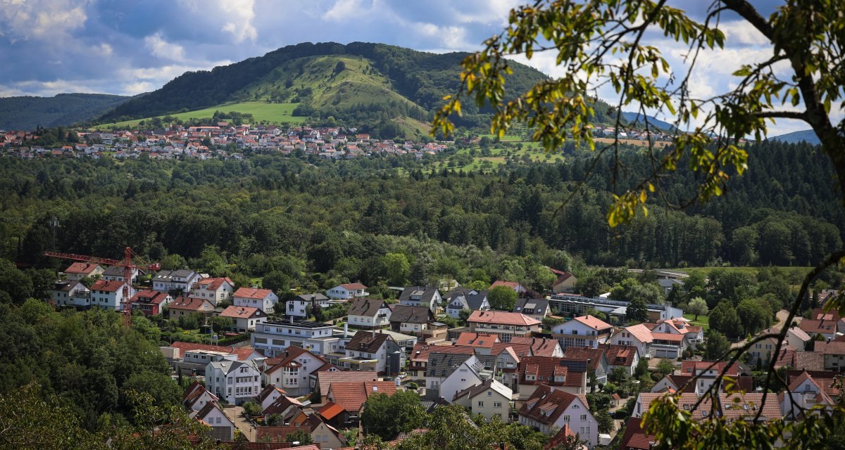 Stadtansicht mit roten Dächern, umgeben von grünen Wäldern und Hügeln im Hintergrund. Ein Baumrahmen im Vordergrund. Stadtansicht mit roten Dächern, umgeben von grünen Wäldern und Hügeln im Hintergrund. Ein Baumrahmen im Vordergrund.