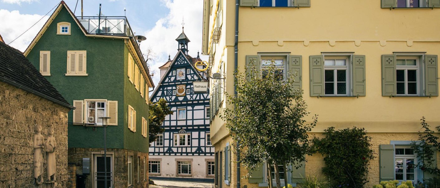 Blick auf ein historische Fachwerkhaus in Bad Überkingen, eingerahmt von bunten Gebäuden, dem Rathaus und einem Baum im Vordergrund., © SMG, Sarah Schmid Blick auf ein historische Fachwerkhaus in Bad Überkingen, eingerahmt von bunten Gebäuden, dem Rathaus und einem Baum im Vordergrund., © SMG, Sarah Schmid