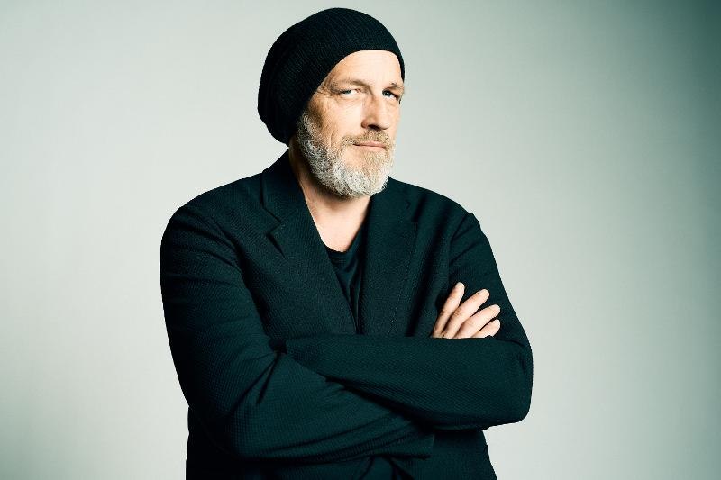 Mann mit Bart und Mütze, in schwarzer Kleidung, verschränkt die Arme vor neutralem Hintergrund., © Roth & Friends