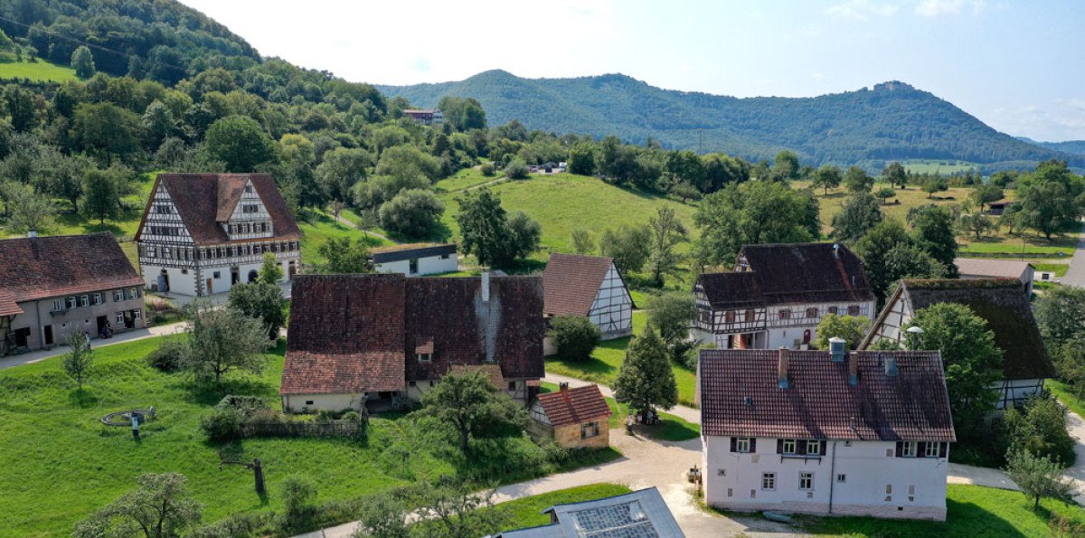 Luftaufnahme eines Dorfes mit traditionellen Fachwerkhäusern, umgeben von grünen Hügeln und Bäumen. Im Hintergrund sind bewaldete Berge zu sehen., © Reiner Enkelmann Luftaufnahme eines Dorfes mit traditionellen Fachwerkhäusern, umgeben von grünen Hügeln und Bäumen. Im Hintergrund sind bewaldete Berge zu sehen., © Reiner Enkelmann