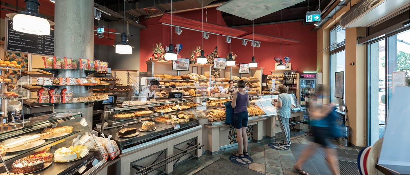 Eine Bäckerei mit Kunden, die an der Theke stehen. Verschiedene Backwaren sind in den Auslagen zu sehen., © Trölsch GmbH