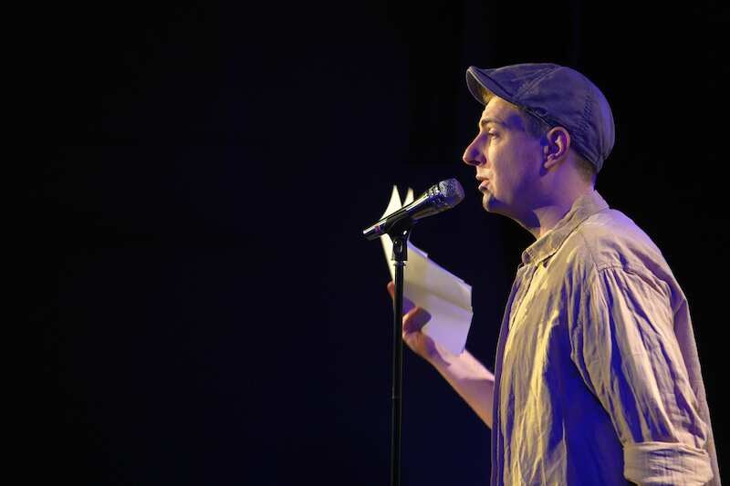Ein Mann mit Mütze spricht in ein Mikrofon und hält ein Blatt Papier. Er steht auf einer Bühne, vermutlich bei einem Poetry Slam., © Kulturhaus Schwanen Ein Mann mit Mütze spricht in ein Mikrofon und hält ein Blatt Papier. Er steht auf einer Bühne, vermutlich bei einem Poetry Slam., © Kulturhaus Schwanen