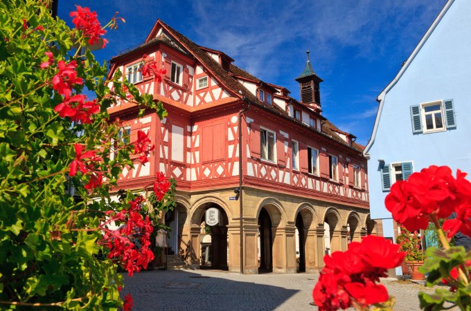 Ein historisches Fachwerkhaus mit roter Fassade und Arkaden. Im Vordergrund blühen rote Blumen, der Himmel ist blau., © WTM GmbH Waiblingen Ein historisches Fachwerkhaus mit roter Fassade und Arkaden. Im Vordergrund blühen rote Blumen, der Himmel ist blau., © WTM GmbH Waiblingen
