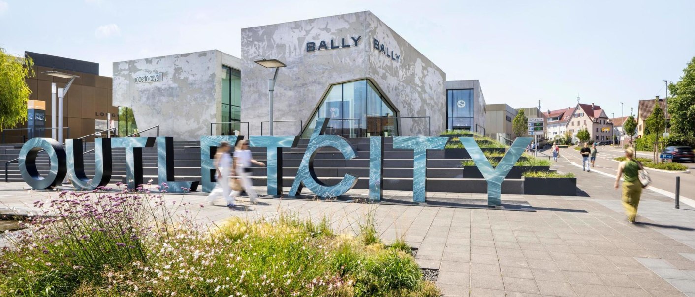 Gro&szlig;e Buchstaben formen das Wort 'OUTLETCITY' vor modernen Geb&auml;uden. Menschen gehen vorbei, w&auml;hrend Blumen im Vordergrund bl&uuml;hen., &copy; OUTLETCITY AG