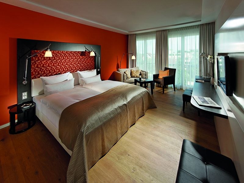 Modernes Hotelzimmer mit großem Bett, oranger Wand, Sofa, Schreibtisch und Fernseher. Helle Vorhänge und Holzboden schaffen eine gemütliche Atmosphäre., © Parkhotel Stuttgart Messe-Airport