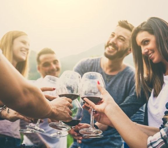Eine Gruppe von Menschen stößt im Freien mit Weingläsern an, lächelnd und fröhlich bei sonnigem Wetter.