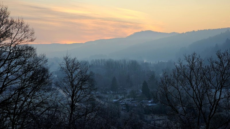 Sonnenuntergang über nebliger Landschaft, Bäume im Vordergrund, Hügel im Hintergrund. Der Himmel ist in warmen Farben gefärbt. Sonnenuntergang über nebliger Landschaft, Bäume im Vordergrund, Hügel im Hintergrund. Der Himmel ist in warmen Farben gefärbt.