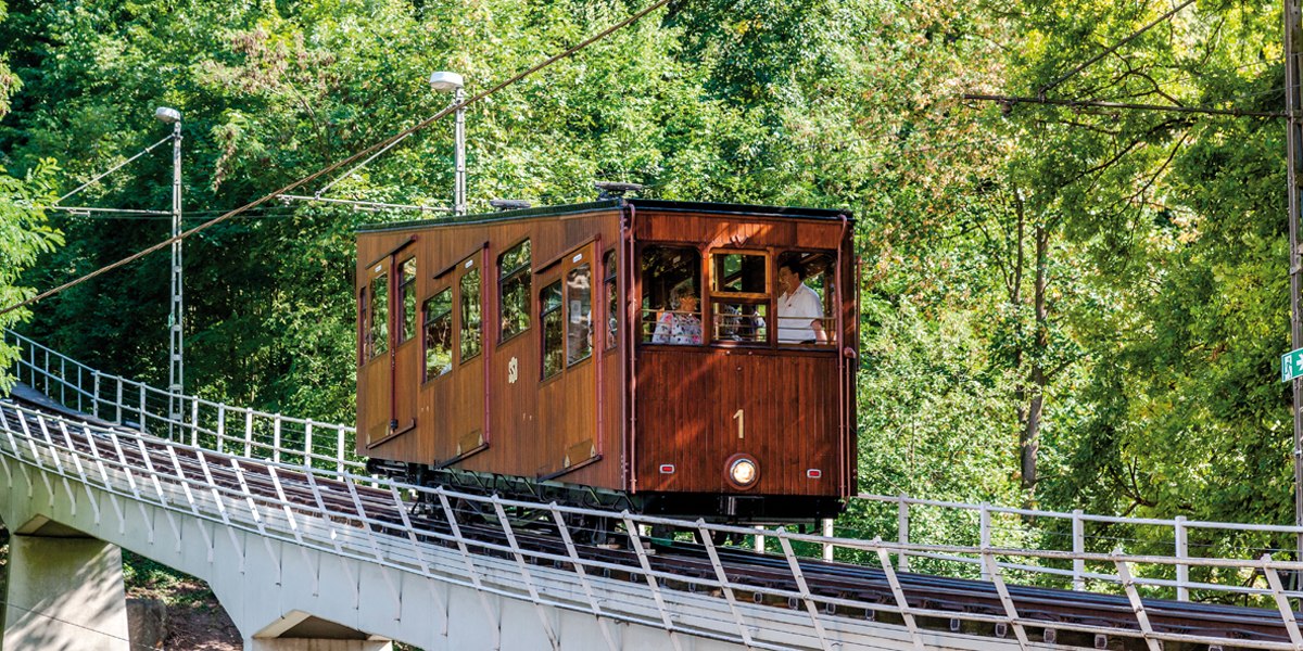 Eine historische Standseilbahn mit Holzverkleidung fährt eine geneigte Strecke entlang, umgeben von dichtem Grün., © TMBW, Gregor Lengler Eine historische Standseilbahn mit Holzverkleidung fährt eine geneigte Strecke entlang, umgeben von dichtem Grün., © TMBW, Gregor Lengler