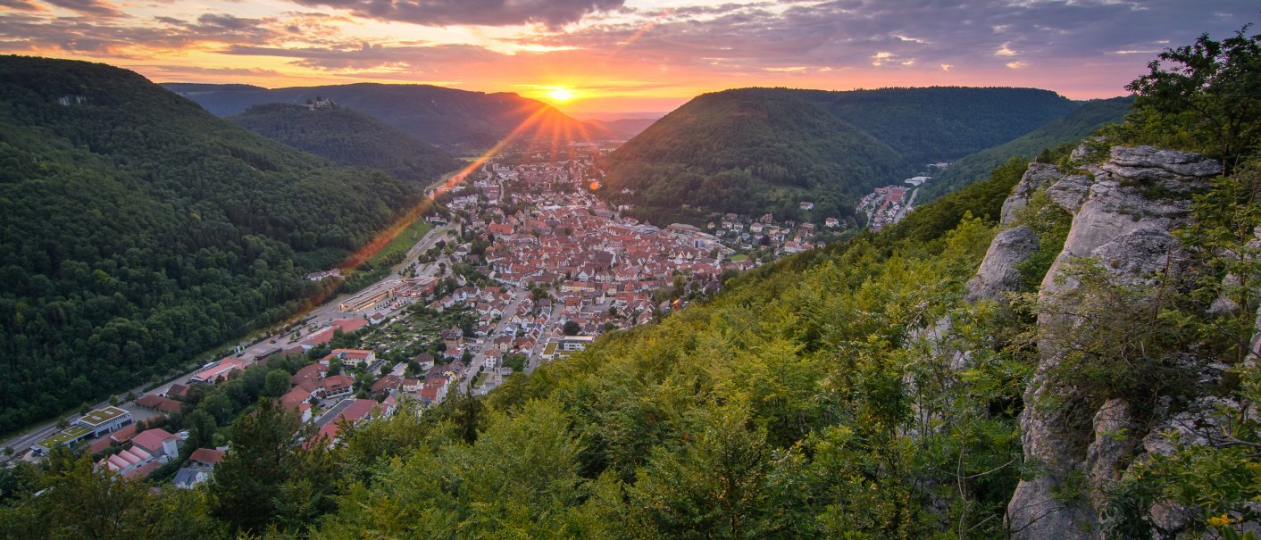 Sonnenuntergang über einer Stadt im Tal, umgeben von grünen Hügeln und Felsen. Der Himmel ist teils bewölkt, die Sonne strahlt orange., © Bad Urach Tourismus Sonnenuntergang über einer Stadt im Tal, umgeben von grünen Hügeln und Felsen. Der Himmel ist teils bewölkt, die Sonne strahlt orange., © Bad Urach Tourismus