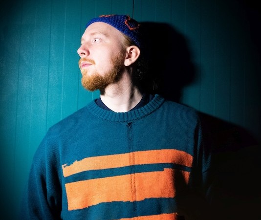 Ein Mann in einem blau-orange gestreiften Pullover steht vor einem dunklen Hintergrund und blickt zur Seite., &copy; Kulturhaus Schwanen