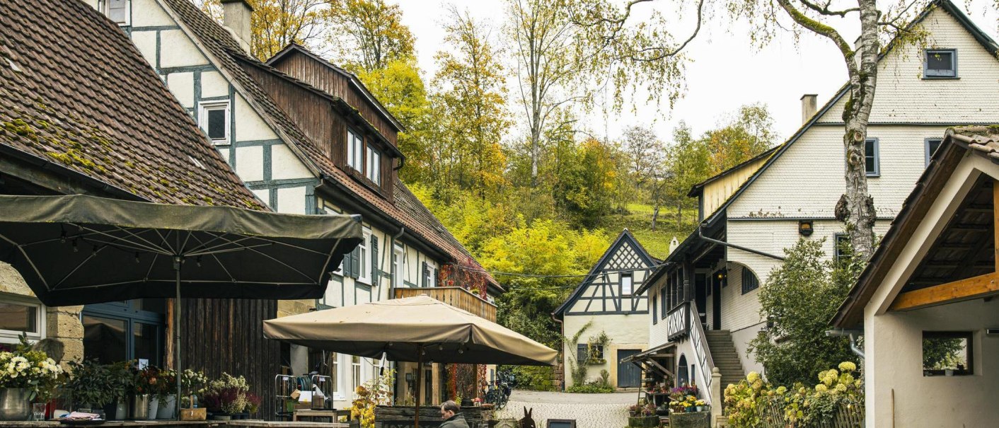 Das Foto zeigt die Eselsmühle. Der Blick der betrachtenden Person folgt der Straße zwischen den Gebäuden hindurch. Links sieht man die Scheune und die Außengastro im Vordergrund, weiter hinten den Eingang zum Hofladen. Auf der rechten Seite ist die Mühle mit Shop., © SMG, Sarah Schmid