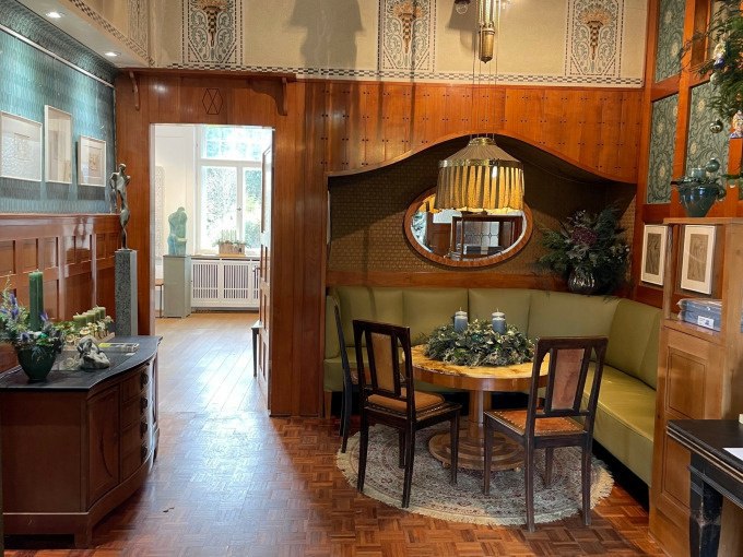 Eleganter Raum mit Holzwänden, runder Tisch mit grüner Eckbank, dekoriert mit Pflanzen und Kunstwerken. Ein Fenster im Hintergrund lässt Licht herein., © Touristik und Marketing GmbH Eleganter Raum mit Holzwänden, runder Tisch mit grüner Eckbank, dekoriert mit Pflanzen und Kunstwerken. Ein Fenster im Hintergrund lässt Licht herein., © Touristik und Marketing GmbH