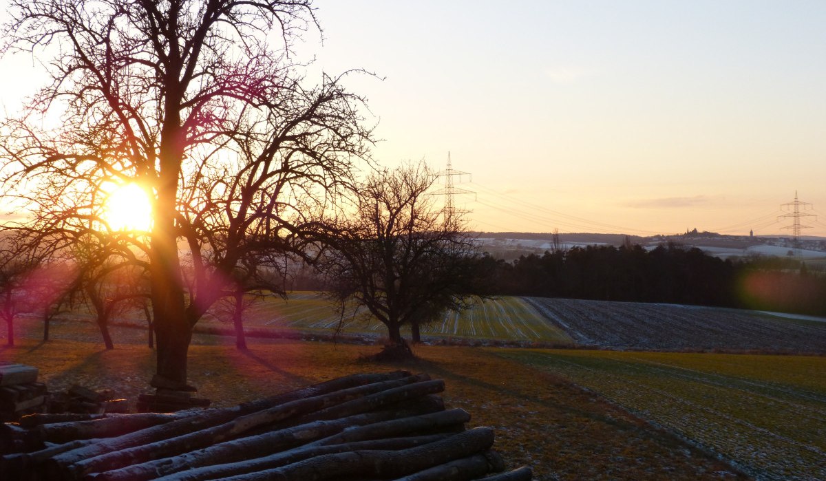 Sonnenuntergang über einem verschneiten Feld mit Bäumen und Holzstapeln im Vordergrund. Strommasten sind am Horizont sichtbar., © Land der 1000 Hügel - Kraichgau-Stromberg Sonnenuntergang über einem verschneiten Feld mit Bäumen und Holzstapeln im Vordergrund. Strommasten sind am Horizont sichtbar., © Land der 1000 Hügel - Kraichgau-Stromberg