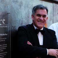 Zwei Männer stehen vor einem Schild des Délice Restaurants. Einer trägt einen Anzug, der andere eine Kochjacke., © Delice