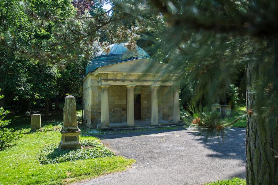 Ein Mausoleum mit Säulen steht in einem grünen Park, umgeben von Bäumen und einem Grabstein. Die Szene wirkt friedlich und historisch., © Copyright: Tourismus & Events Ludwigsburg, Fotograf: Benjamin Stollenberg