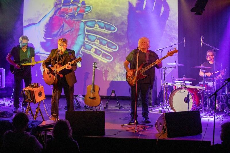 Eine Band performt auf einer B&uuml;hne mit Gitarren, Bass und Schlagzeug. Im Hintergrund sind bunte Projektionen von H&auml;nden zu sehen., &copy; Interessengemeinschaft Kultur Sindelfingen / B&ouml;blingen e.V.