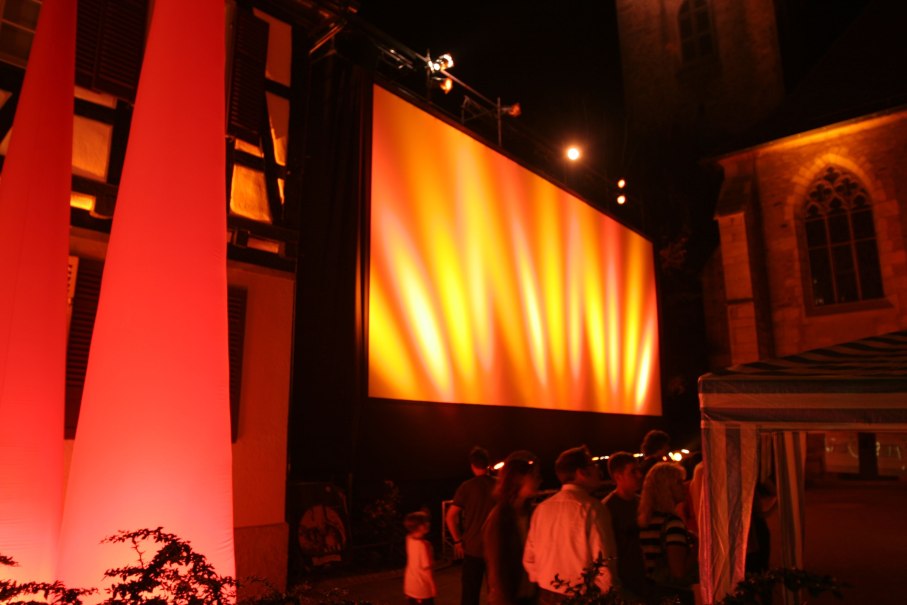 Freiluftkino bei Nacht mit großer, beleuchteter Leinwand. Menschen stehen davor, umgeben von warmem Licht und Gebäuden im Hintergrund., © Torsten Wenzler