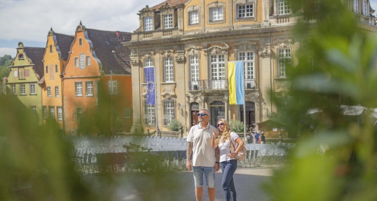 Ein Paar auf dem Marktplatz in Schw&auml;bisch Hall, umgeben von historischen Geb&auml;uden und Fahnen. Sonniges Wetter und gr&uuml;ne Pflanzen im Vordergrund., &copy; Hohenlohe + Schw&auml;bisch Hall Tourismus e. V.