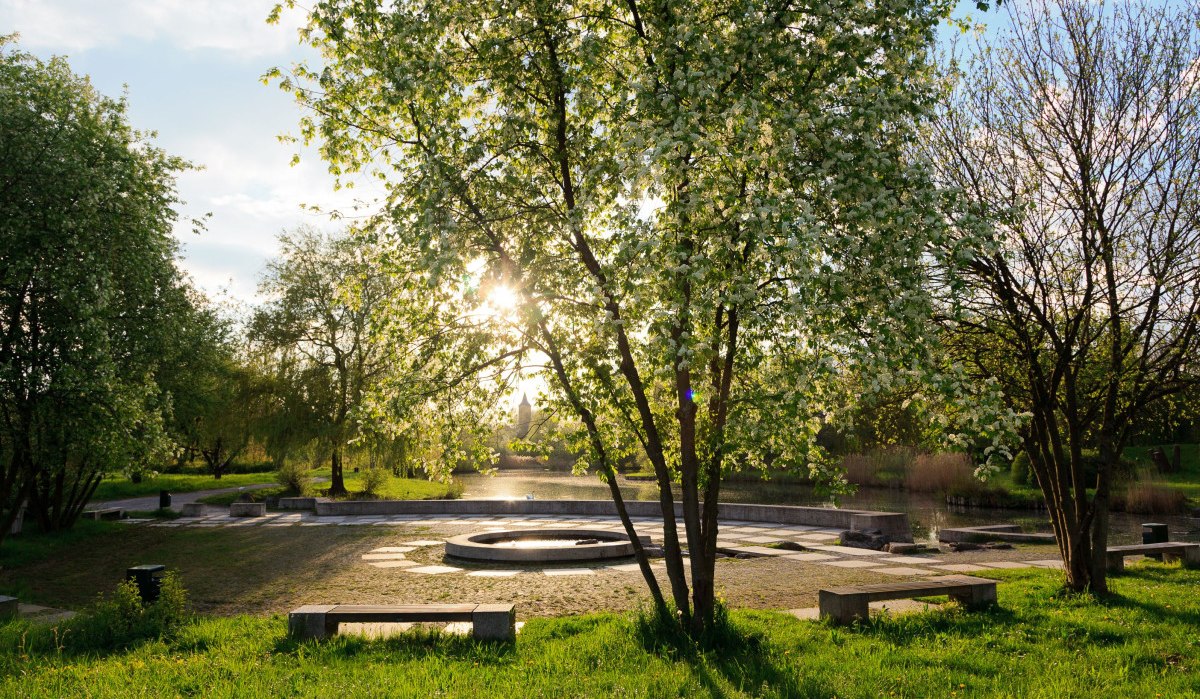Ein Park mit blühenden Bäumen, Bänken und einem runden Brunnen. Die Sonne scheint durch die Blätter, und der Himmel ist leicht bewölkt., © Remstal Tourismus e.V.