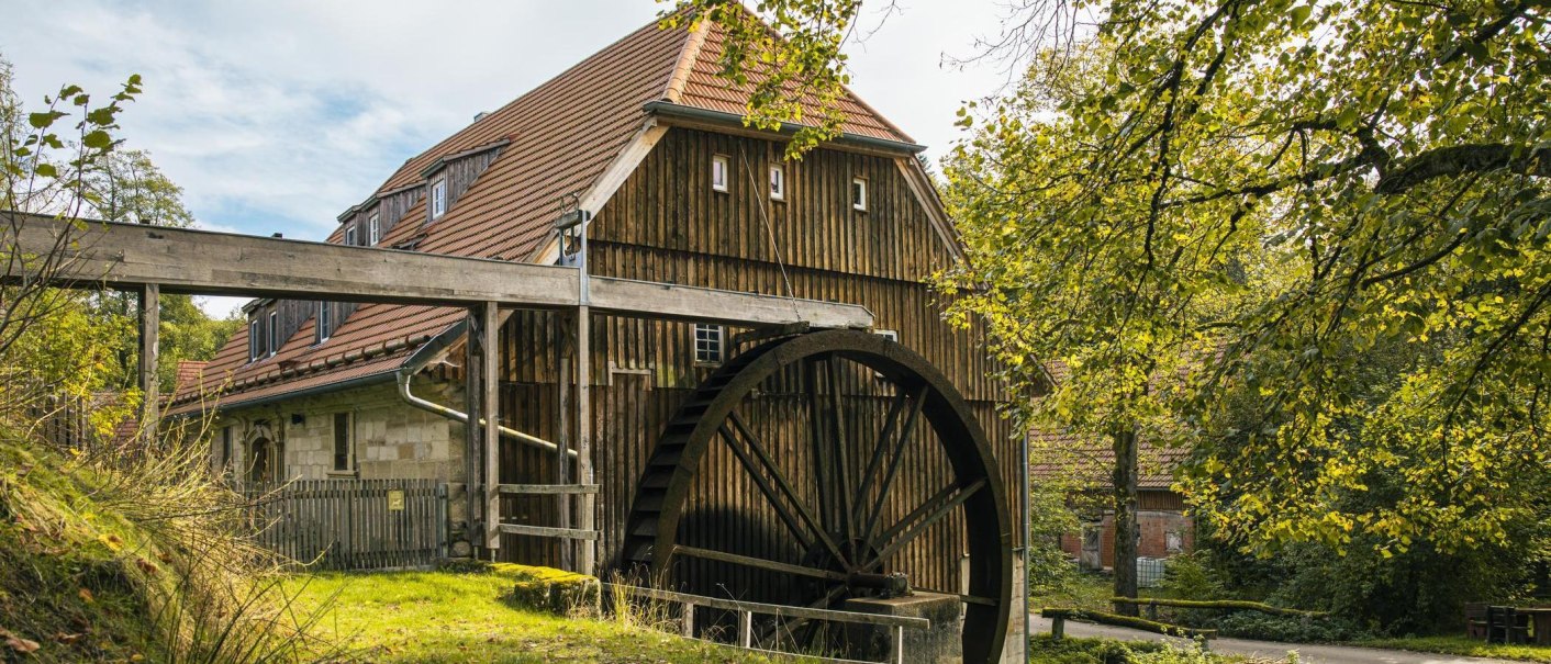 Eine historische Wasserm&uuml;hle mit gro&szlig;em Wasserrad und Fachwerkgeb&auml;ude, umgeben von gr&uuml;ner Natur und B&auml;umen., &copy; SMG, Sarah Schmid
