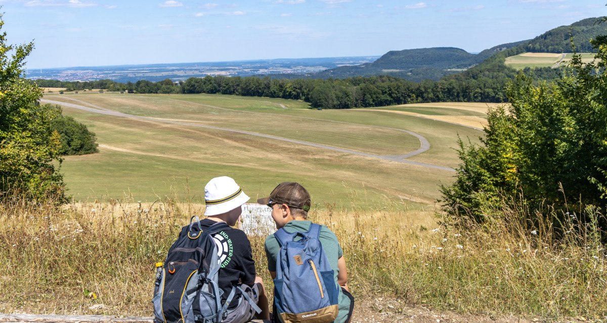 Zwei Personen mit Rucksäcken sitzen auf einem Baumstamm und blicken auf die Landebahn des Segelflugplatzes Hornberg in einer grünen, hügeligen Landschaft., © Foto Thomas Zehnder Zwei Personen mit Rucksäcken sitzen auf einem Baumstamm und blicken auf die Landebahn des Segelflugplatzes Hornberg in einer grünen, hügeligen Landschaft., © Foto Thomas Zehnder
