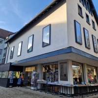 Die Buchhandlung Zimmermann zeigt eine moderne Fassade mit großen Fenstern. Vor dem Eingang stehen Tische mit Büchern zum Verkauf., © cStadtNuertingen Die Buchhandlung Zimmermann zeigt eine moderne Fassade mit großen Fenstern. Vor dem Eingang stehen Tische mit Büchern zum Verkauf., © cStadtNuertingen