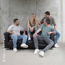 Vier Personen sitzen auf einem schwarzen Sofa vor einer Betonwand und lachen miteinander., &copy; links im Bild