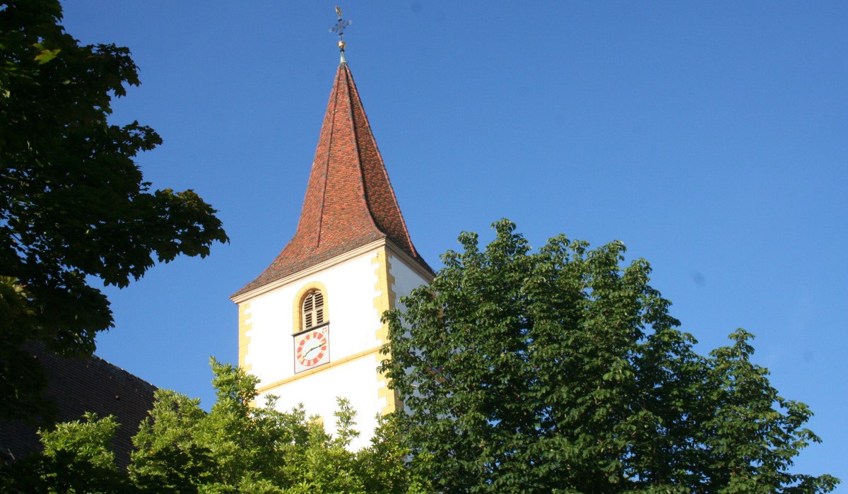 Ein Kirchturm mit rotem Dach und Uhr in Holzgerlingen, umgeben von grünen Bäumen, vor klarem blauem Himmel., © Natur.Nah. Schönbuch & Heckengäu Ein Kirchturm mit rotem Dach und Uhr in Holzgerlingen, umgeben von grünen Bäumen, vor klarem blauem Himmel., © Natur.Nah. Schönbuch & Heckengäu