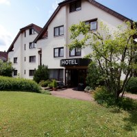 Ein Hotelgebäude mit weißer Fassade und einem Schild "HOTEL" über dem Eingang, umgeben von gepflegtem Garten und Bäumen., © Hotel Flora