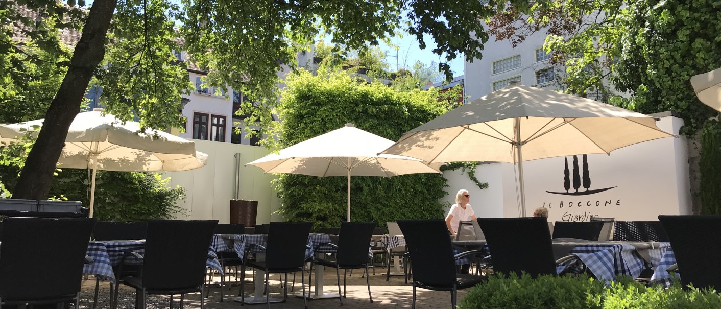 Ein sonniger Garten mit Tischen, St&uuml;hlen und Sonnenschirmen, umgeben von B&auml;umen und Pflanzen. Im Hintergrund ist eine Wand mit der Aufschrift 'Il Boccone' sichtbar., &copy; Restaurant Il Boccone
