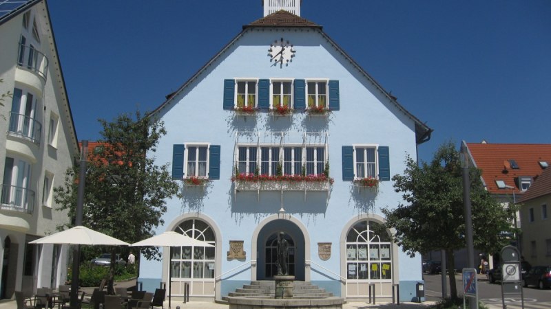 Blaues Gebäude mit Uhr und Blumenkästen, davor ein Brunnen und Café mit Sonnenschirmen, bei sonnigem Wetter., © http://www.rutesheim.de/servlet/PB/menu/1110073/index.html