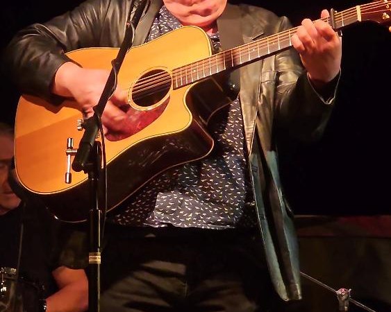Ein Gitarrist mit Brille und Lederjacke spielt konzentriert auf der B&uuml;hne, w&auml;hrend ein Schlagzeuger im Hintergrund zu sehen ist., &copy; Foto Bernd Hoffmann