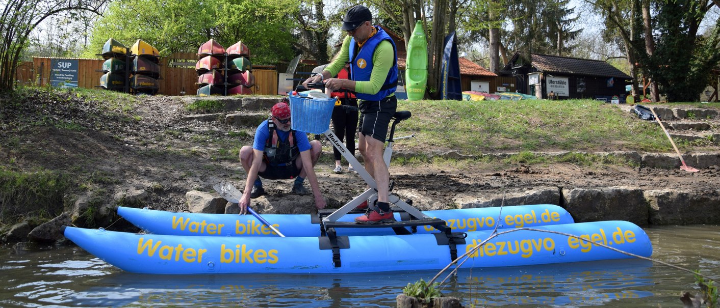Zwei Personen mit Schwimmwesten bereiten ein Wasserfahrrad am Ufer vor. Im Hintergrund sind Kanus und ein Schild für Stand-Up-Paddling zu sehen., © Die Zugvögel