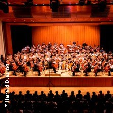 Ein großes Cello-Orchester spielt auf einer Bühne in einem Konzertsaal vor Publikum., © links im Bild Ein großes Cello-Orchester spielt auf einer Bühne in einem Konzertsaal vor Publikum., © links im Bild