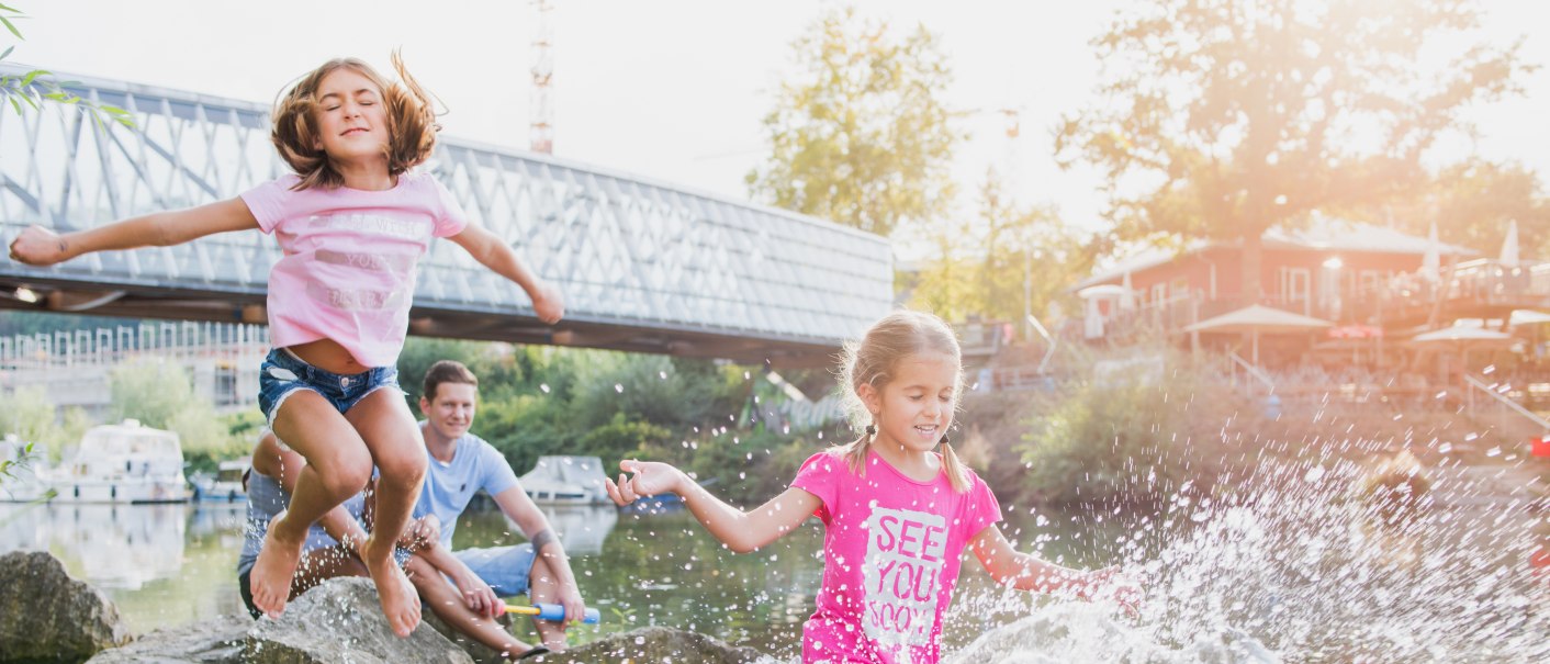 Kinder springen fröhlich von Steinen ins Wasser am Neckarstrand in Remseck. Eine Brücke und Boote sind im Hintergrund zu sehen., © Bebop Media