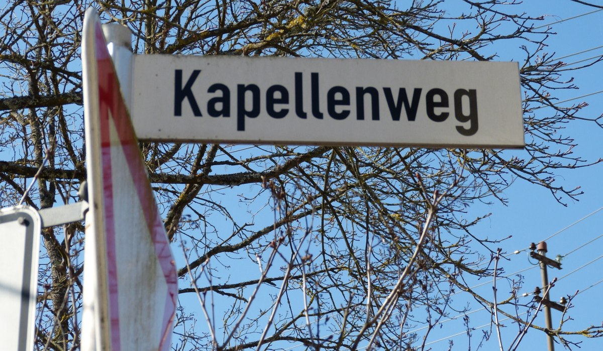 Ein Stra&szlig;enschild mit der Aufschrift 'Kapellenweg' vor einem blauen Himmel und kahlen &Auml;sten., &copy; Natur.Nah. Sch&ouml;nbuch & Heckeng&auml;u