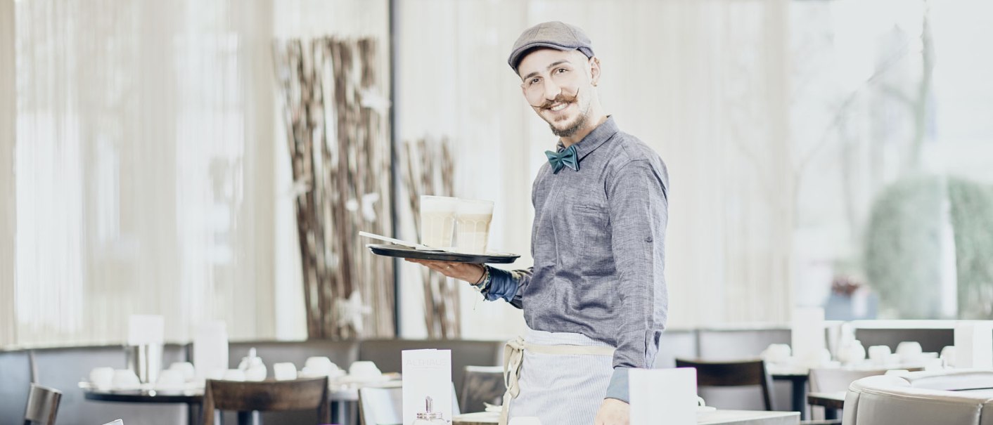 Ein lächelnder Kellner mit Mütze und Fliege serviert Getränke in einem hellen, modernen Frühstücksbereich., © MASEVEN Stuttgart SI-Centrum Ein lächelnder Kellner mit Mütze und Fliege serviert Getränke in einem hellen, modernen Frühstücksbereich., © MASEVEN Stuttgart SI-Centrum