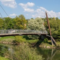 Eine moderne Fußgängerbrücke überspannt einen Fluss, umgeben von grüner Vegetation. Im Hintergrund ist ein Kirchturm zu sehen., © Stuttgart-Marketing GmbH Eine moderne Fußgängerbrücke überspannt einen Fluss, umgeben von grüner Vegetation. Im Hintergrund ist ein Kirchturm zu sehen., © Stuttgart-Marketing GmbH
