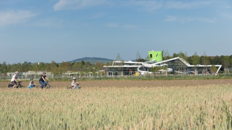 Familie radelt durch ein Feld, modernes Gebäude mit Rutschen im Hintergrund, blauer Himmel., © Stadt Fellbach Familie radelt durch ein Feld, modernes Gebäude mit Rutschen im Hintergrund, blauer Himmel., © Stadt Fellbach