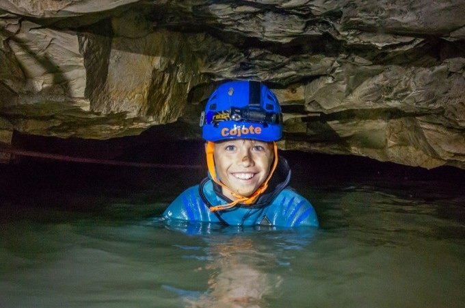 Eine Person mit Helm und Stirnlampe steht lächelnd in einer wassergefüllten Höhle. Die Umgebung ist von Felsen umgeben., © SwabianTravel Eine Person mit Helm und Stirnlampe steht lächelnd in einer wassergefüllten Höhle. Die Umgebung ist von Felsen umgeben., © SwabianTravel
