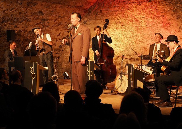 Jazzband spielt in rustikalem Keller. Sänger im Anzug, Musiker mit Saxophon, Kontrabass, Klavier, Schlagzeug und Gitarre. Publikum im Vordergrund., © Theater im Schlosskeller Jazzband spielt in rustikalem Keller. Sänger im Anzug, Musiker mit Saxophon, Kontrabass, Klavier, Schlagzeug und Gitarre. Publikum im Vordergrund., © Theater im Schlosskeller