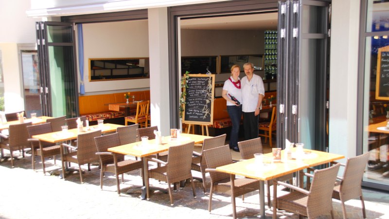 Ein Restaurant mit offenen Türen, draußen stehen Tische und Stühle. Zwei Personen stehen im Eingang neben einer Tafel mit Menü. Ein Restaurant mit offenen Türen, draußen stehen Tische und Stühle. Zwei Personen stehen im Eingang neben einer Tafel mit Menü.