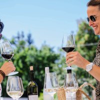 Zwei Frauen genie&szlig;en Wein im Freien, sto&szlig;en mit Gl&auml;sern an. Im Hintergrund sind B&auml;ume und ein blauer Himmel zu sehen., &copy; SMG, Thomas Niederm&uuml;ller