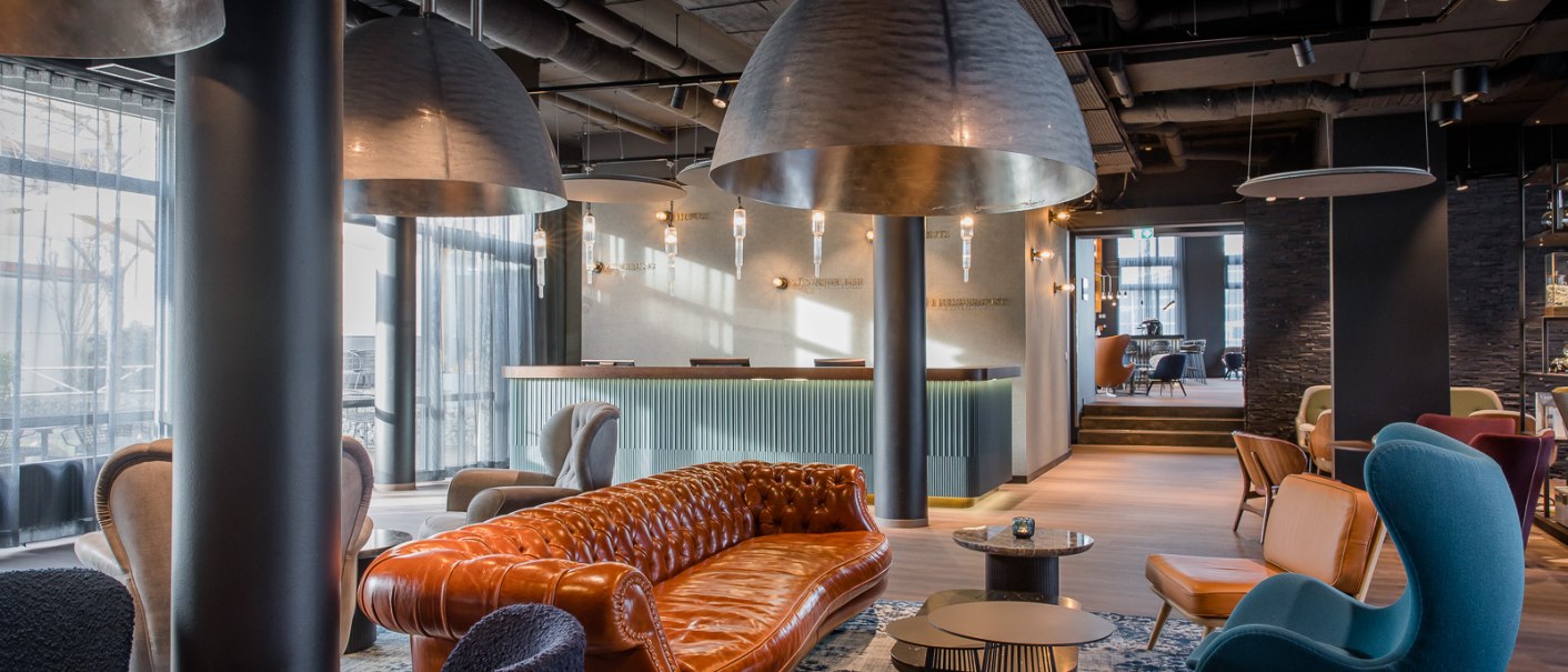 Elegante Hotellobby mit braunem Ledersofa, blauen und grauen Sesseln, großen Hängelampen und einem Empfangstresen im Hintergrund., © TOMAS Elegante Hotellobby mit braunem Ledersofa, blauen und grauen Sesseln, großen Hängelampen und einem Empfangstresen im Hintergrund., © TOMAS