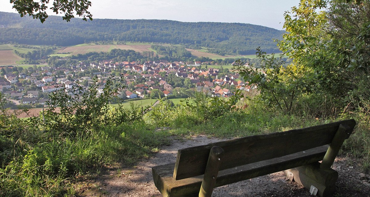Eine Holzbank bietet einen malerischen Blick auf das Dorf Gündelbach, umgeben von grünen Feldern und bewaldeten Hügeln., © Land der 1000 Hügel - Kraichgau-Stromberg Eine Holzbank bietet einen malerischen Blick auf das Dorf Gündelbach, umgeben von grünen Feldern und bewaldeten Hügeln., © Land der 1000 Hügel - Kraichgau-Stromberg