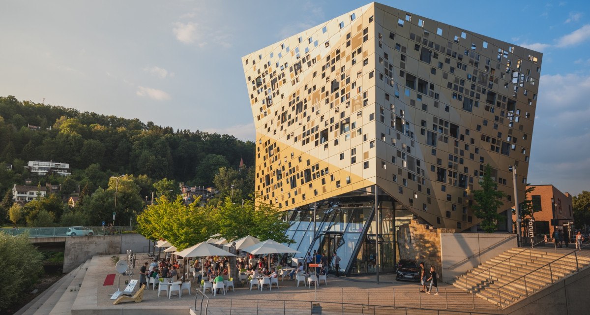 Modernes Gebäude mit goldener Fassade, viele Fenster. Menschen sitzen unter Sonnenschirmen. Umgeben von Bäumen und Treppen., © Remstal Tourismus e.V. I Architekt: isin Architekten