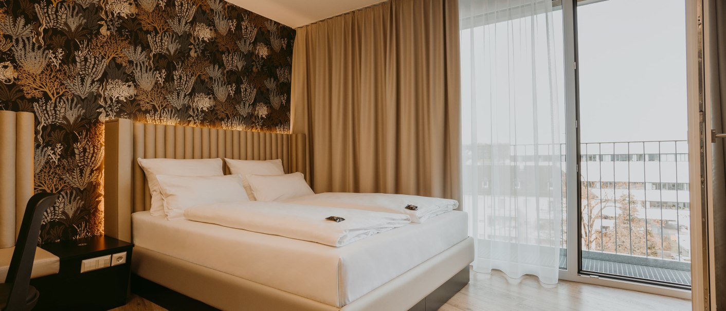 Modernes Hotelzimmer mit Doppelbett, eleganter Tapete und Zugang zu einem Balkon. Helle Vorhänge und Holzboden ergänzen das Design., © TOMAS Modernes Hotelzimmer mit Doppelbett, eleganter Tapete und Zugang zu einem Balkon. Helle Vorhänge und Holzboden ergänzen das Design., © TOMAS