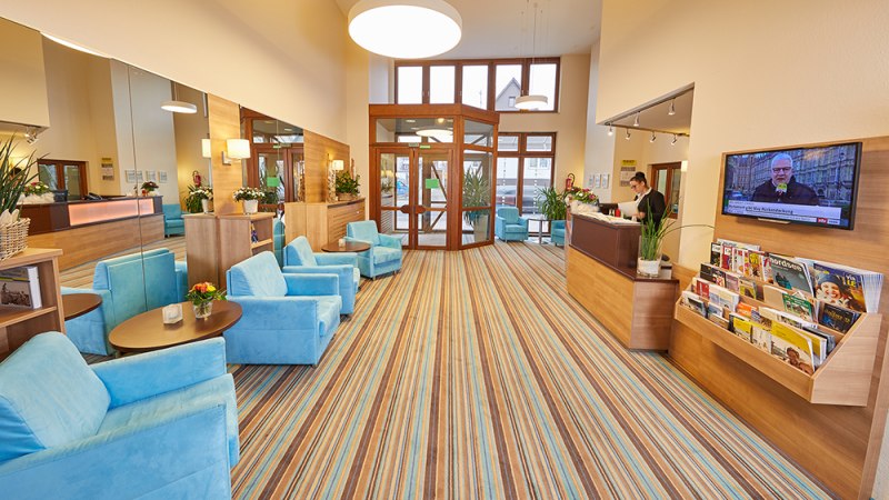 Helle Lobby mit blauen Sesseln, Holzmöbeln und gestreiftem Teppich. Rezeption mit Mitarbeiter, TV an der Wand, Zeitschriftenständer., © TOMAS